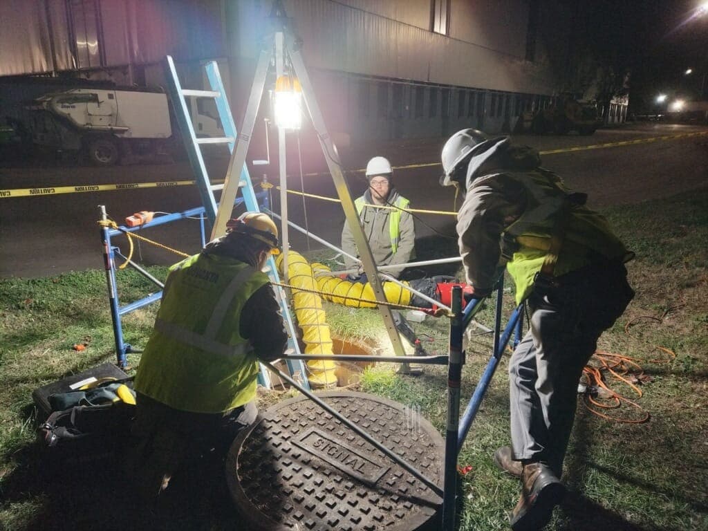 Electrisource crew performing planned confined-space work at a manhole with retrieval equipment, ventilation, and lighting in place
